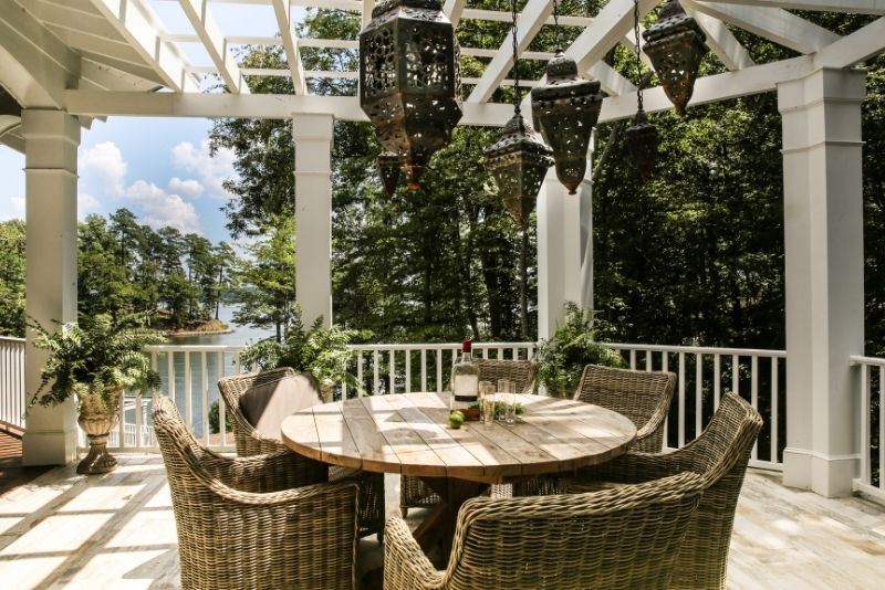 White pergola with hanging lanterns over a wicker outdoor dining set and lake views